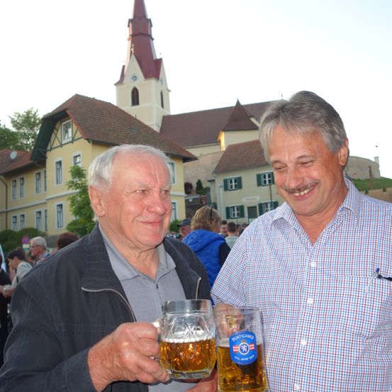 Zwei ältere Männer stehen vor einer Kirche und halten Gläser mit Bier in den Händen. Sie lächeln und posieren für ein Foto. Hinter ihnen ist eine Menschenmenge zu sehen, und einige stehen unter Regenschirmen.