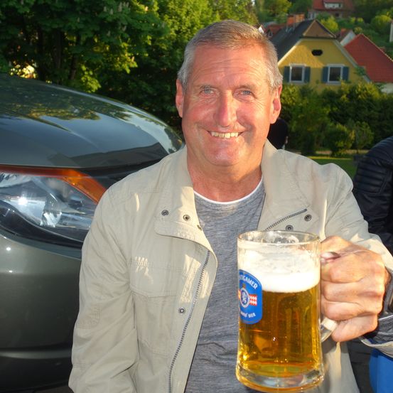 Ein Mann in einer hellbeigen Jacke hält vor einem Auto ein Glas Bier in der Hand und lächelt.