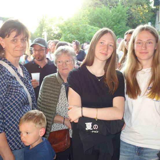 Eine Familie und Freunde sind draußen versammelt. Eine Frau links trägt ein blaues Blumenhemd, und ein Junge neben ihr schaut zur Seite. Eine andere Frau mit Brille hält eine braune Tasche. Eine Frau in Schwarz hat eine Halskette, und ein junges Mädchen neben ihr trägt ein weißes Shirt.