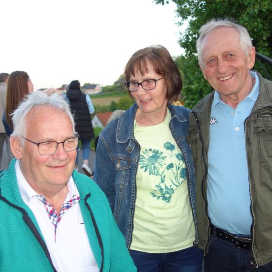 Drei ältere Menschen stehen vor einem Baum. Sie alle lächeln. Die Frau trägt eine Brille und eine Jeansjacke. Der Mann rechts trägt eine Jacke. Im Hintergrund sind einige Menschen zu sehen, die gehen.