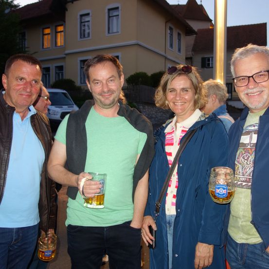 Vier Personen, drei Männer und eine Frau, stehen eng beieinander, lächeln und halten Bierkrüge. Sie sind draußen, mit einem Gebäude und Bäumen im Hintergrund.