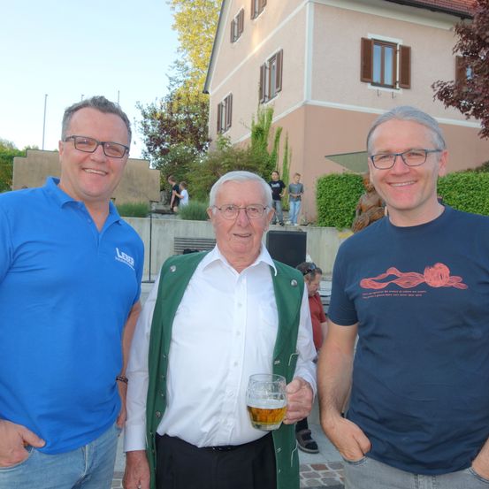 Drei Männer stehen vor einem Gebäude. Der Mann auf der linken Seite trägt ein blaues Poloshirt mit Logo, der Mann in der Mitte trägt eine grüne Weste über einem weißen Hemd und hält ein Glas Bier in der Hand, und der Mann auf der rechten Seite trägt ein dunkelblaues T-Shirt. Hinter ihnen ist ein Haus mit mehreren Fenstern und einer Statue zu sehen.