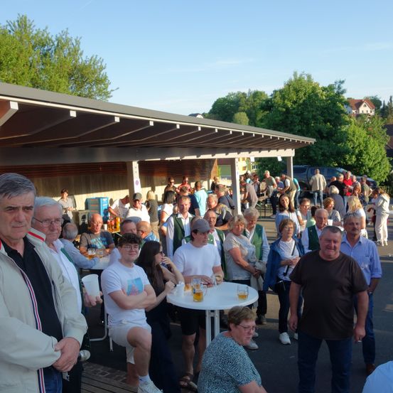 Eine große Gruppe von Menschen steht und sitzt um einen weißen Tisch im Freien unter einem Dach.