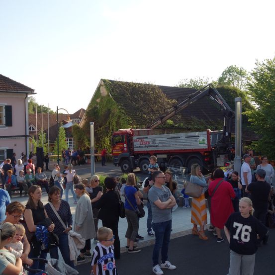 Eine Menschenmenge steht auf einem Freigelände. Eine Person hält ein Baby, und ein Mann schaut auf einen Kran. Ein Lastwagen ist vor dem Gebäude geparkt. Das Gebäude hat ein moosbewachsenes Dach und einige Fenster.