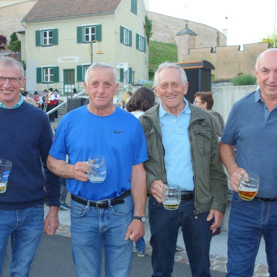 Vier Männer stehen vor einem Gebäude mit einem Schild, das 'Pfarrheim' steht. Sie lächeln und halten Gläser mit Bier.