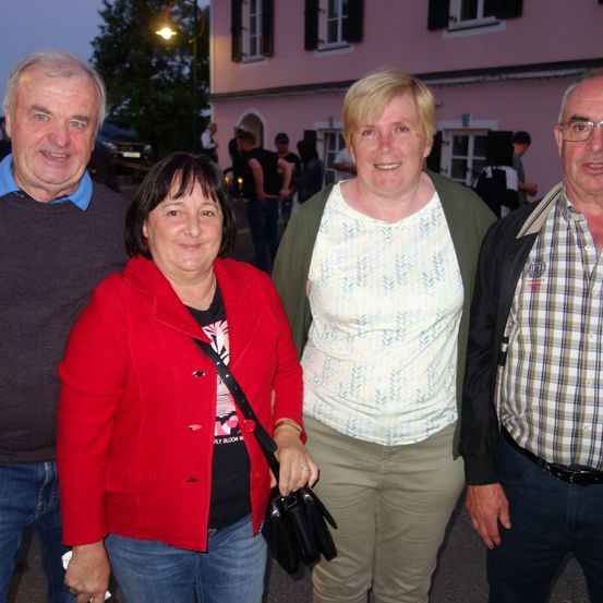 Vier Menschen stehen bei Nacht draußen. Zwei Frauen lächeln für ein Foto. Die Frau auf der linken Seite trägt eine rote Jacke und die Frau auf der rechten Seite ein weißes Oberteil. Die beiden Männer an den Seiten lächeln. Dahinter sind Menschen und ein rosa Gebäude.