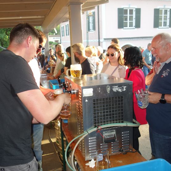 Ein Mann zapft Bier aus einem Zapfhahn in ein Glas, während andere stehen und ihre Getränke genießen.