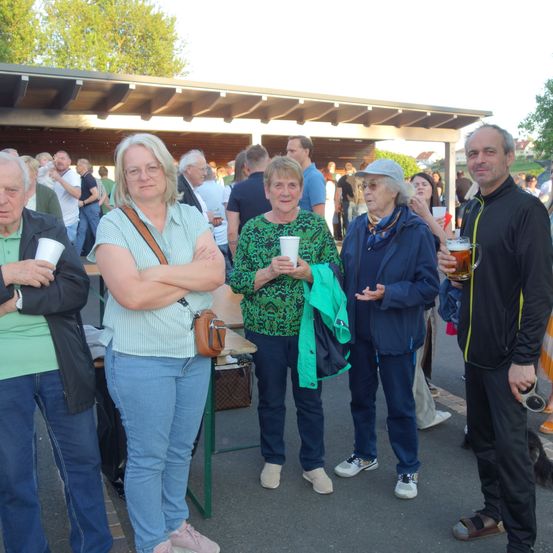 Fünf Personen stehen in einer Gruppe draußen, einige mit Getränken in der Hand. Eine Frau links hält einen Becher in der Hand. Eine Frau in der Mitte hält einen Becher in beiden Händen. Eine Frau rechts hält ein Glas Bier.