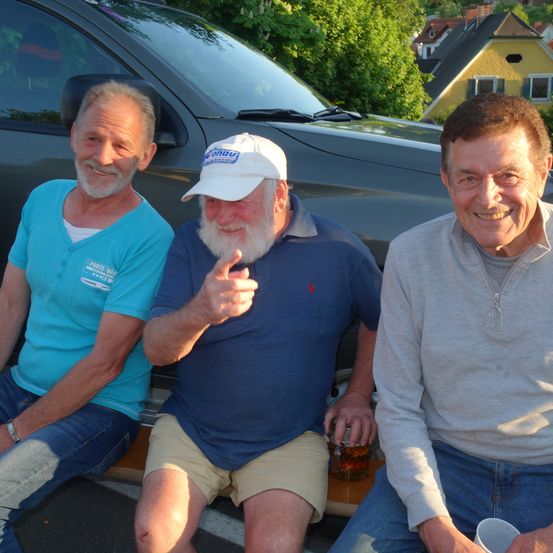 Drei Männer sitzen neben einem schwarzen Auto, mit Bäumen und einem Haus im Hintergrund. Ein Mann gibt einen Daumen hoch, ein anderer hält ein Glas mit einem Getränk und der dritte lächelt.