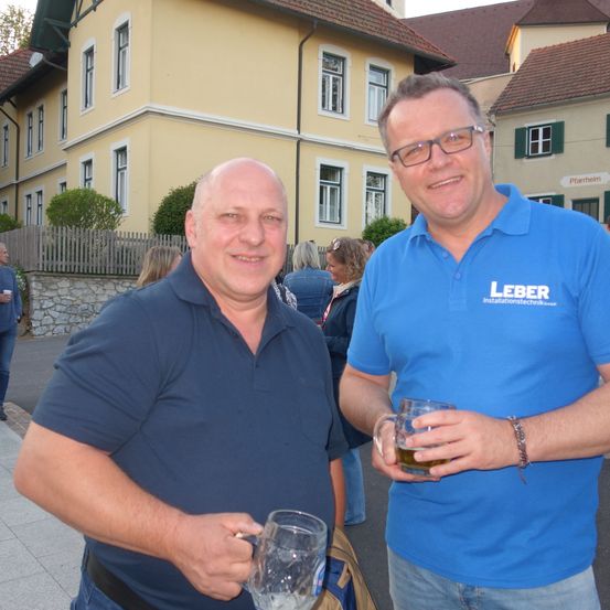 Zwei Männer stehen vor einem Gebäude. Einer von ihnen hält ein Glas Bier. Er trägt ein blaues Poloshirt mit dem Wort 'Leber' darauf. Der andere Mann trägt ein blaues Poloshirt und hält ein Glas Bier. Sie lächeln und scheinen für ein Foto zu posieren.