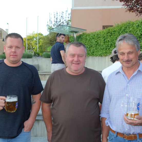 Drei Männer stehen draußen mit Getränken in den Händen. Einer hält ein Bierglas, der andere ein Bierkrug. Der Mann in der Mitte trägt ein braunes T-Shirt.