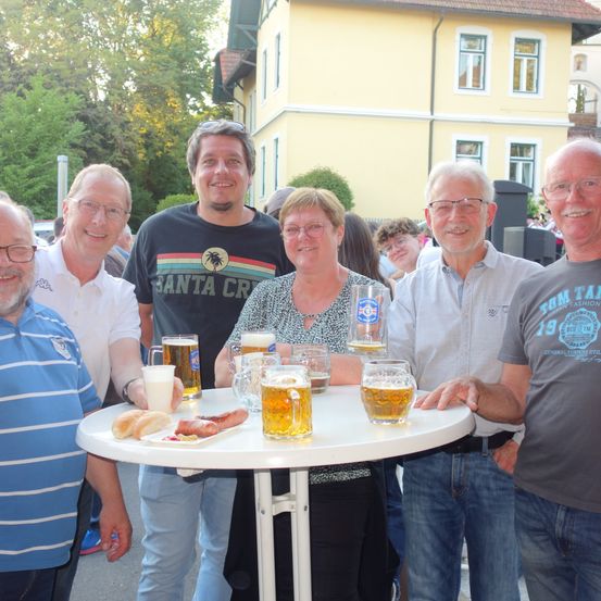 Eine Gruppe von fünf Personen lächelt und posiert für ein Foto vor einem Gebäude. Sie halten Biergläser und eine Frau hält einen Tablett mit Essen.