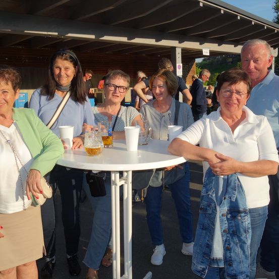 Eine Gruppe lächelnder Menschen steht an einem sonnigen Tag um einen Tisch mit Biergläsern.