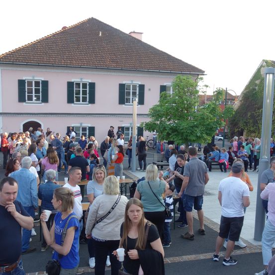 Eine Menschenmenge, bestehend aus Männern und Frauen, versammelt sich vor einem rosafarbenen Gebäude. Einige sitzen auf Bänken, andere stehen, und sie tragen verschiedene Arten von Kleidung.