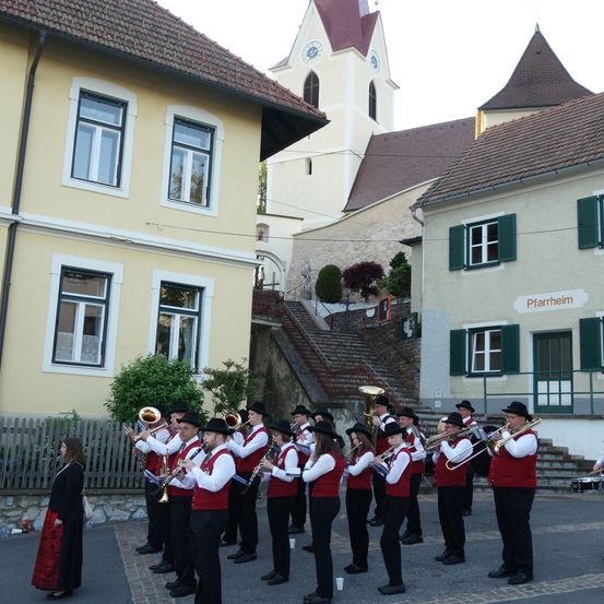 Eine Blechblaskapelle spielt vor einem gelben Gebäude, mit einem Kirchturm im Hintergrund.