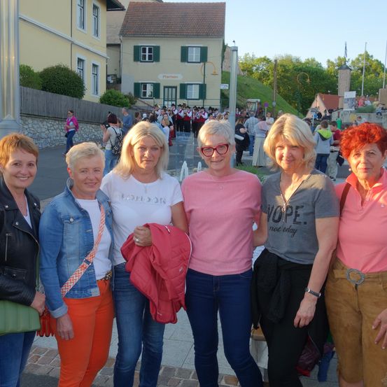 Fünf Frauen in unterschiedlich gefärbter Kleidung stehen vor einem Gebäude mit einer Menschenmenge und einer Marschkapelle im Hintergrund.