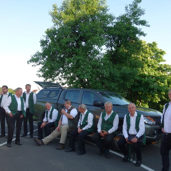 Eine Gruppe von Männern in grünen Westen und weißen Hemden posiert für ein Foto auf einem Parkplatz.