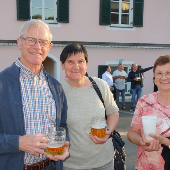 Drei Erwachsene lächeln und halten Getränke in der Hand. Eine Frau in der Mitte mit kurzen Haaren und ein Mann links von ihr mit Brille. Eine Frau rechts mit einem weißen Becher. Dahinter steht eine Gruppe von Menschen vor einem rosa Gebäude mit grünen Fensterläden.