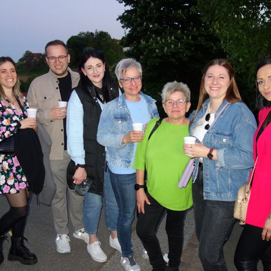 Eine Gruppe von Menschen steht bei Nacht draußen. Sie lächeln und halten Tassen. Sie sind lässig in verschiedenen Farben und Stilen gekleidet.