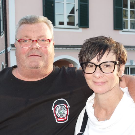 Ein Mann und eine Frau stehen vor einem Gebäude. Der Mann trägt ein schwarzes T-Shirt mit Logo und roten Brillengläsern. Die Frau trägt ein weißes Hemd, eine Brille und eine Halskette.