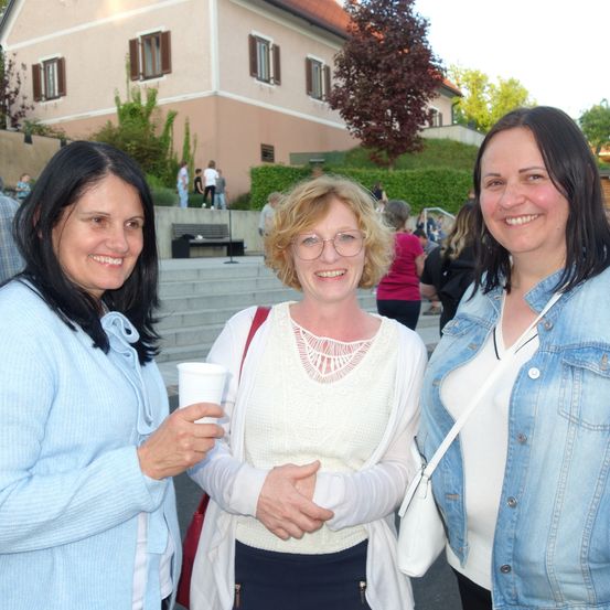 Drei Frauen stehen vor einem Gebäude mit Treppen und Grünanlagen und lächeln in die Kamera. Die Frau links hält einen Becher, die mittlere trägt eine Brille und eine rote Umhängetasche, und die Frau rechts trägt eine Jeansjacke.