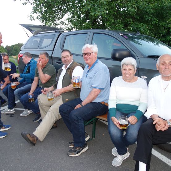 Eine Gruppe älterer Männer und Frauen sitzt auf einer Bank vor einem geparkten Fahrzeug. Sie halten alle Gläser mit Bier in den Händen.