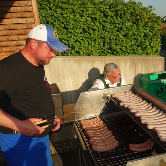 Ein Mann grillt Würste auf einem Grill im Freien, während ein anderer Mann zusieht. Dahinter befindet sich eine Betonmauer mit einem Mülleimer und einem Busch.