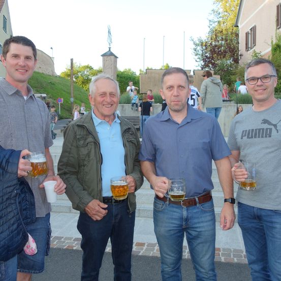 Vier Männer stehen auf einer Treppe und halten Biergläser in der Hand. Sie lächeln und scheinen für ein Foto zu posieren. Hinter ihnen befinden sich Treppen, ein Grashügel und Gebäude.