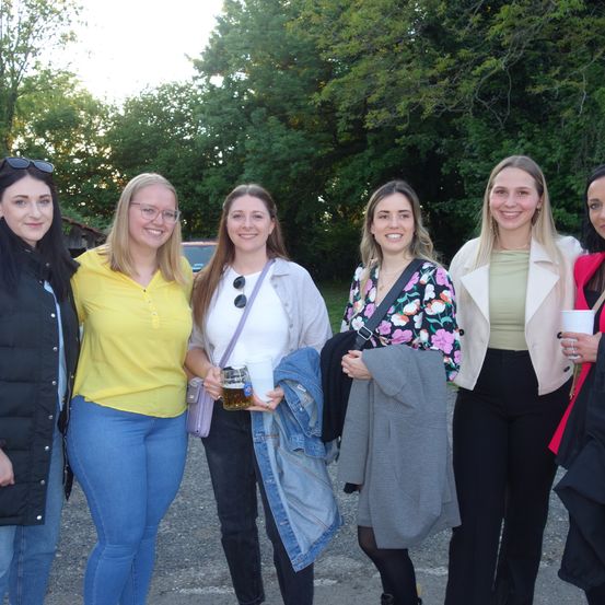 Fünf Frauen stehen zusammen auf einem Parkplatz mit Bäumen im Hintergrund. Eine Frau hält einen Becher Bier, während die anderen lächeln.