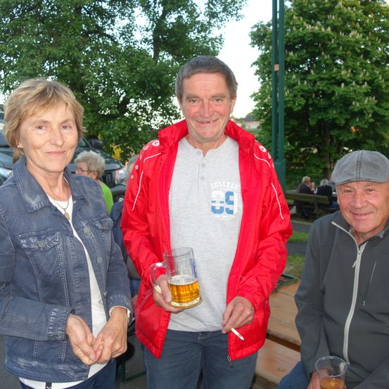Drei ältere Menschen lächeln und stehen in einem Außenbereich. Der Mann in der Mitte hält ein Bierglas und eine Zigarette. Die Frau links trägt eine Jeansjacke und eine Halskette. Der Mann rechts trägt eine graue Jacke und einen Hut.