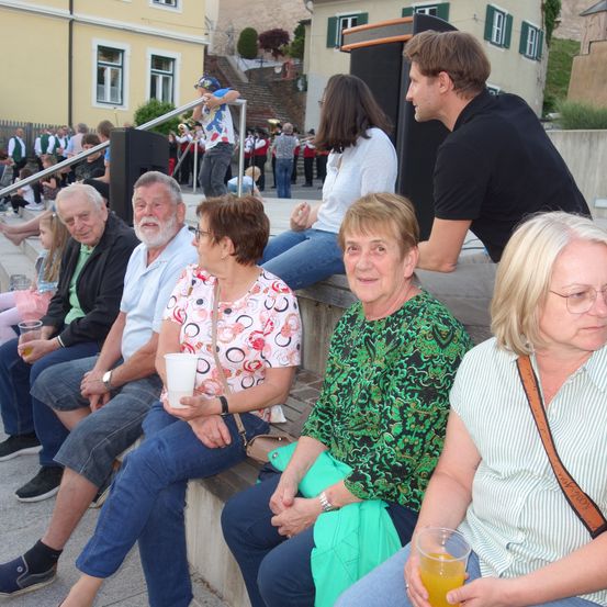 Eine Gruppe älterer Menschen sitzt zusammen auf einer Betontreppe. Ein Mann mit schwarzem Shirt steht hinter ihnen. Sie beobachten wahrscheinlich ein Konzert.