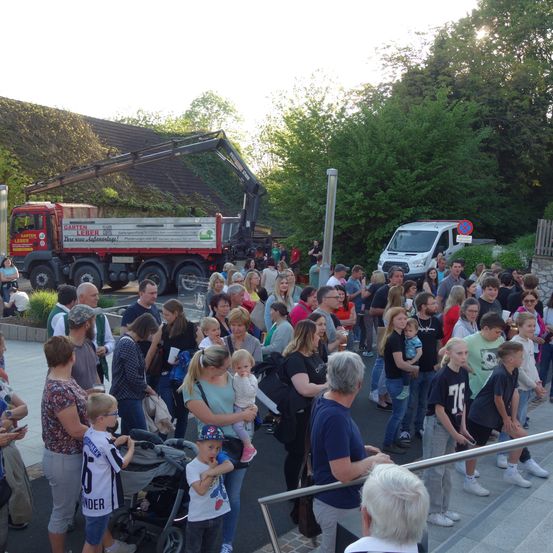 Eine große Menschenmenge, darunter Familien mit kleinen Kindern, versammelt sich in einem öffentlichen Bereich. Ein roter Lastwagen mit der Aufschrift 'Garden Labor' ist in der Nähe geparkt, und ein weißer Van befindet sich im Hintergrund. Die Menschen stehen und unterhalten sich, einige halten Tassen. Das Gebiet ist von Grün und Bäumen umgeben.