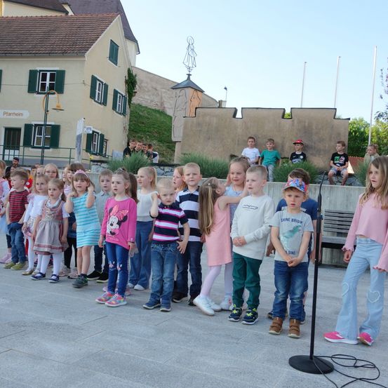 Eine Gruppe junger Kinder, möglicherweise in einem Chor, steht vor einem Mikrofon, während einige Erwachsene auf einer Bank im Hintergrund sitzen.