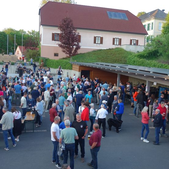 Eine große Menschenmenge steht und unterhält sich im Freien. Einige von ihnen halten Tassen. Es gibt Stände an der Seite, vor denen Menschen stehen. Hinter der Menge befindet sich ein Haus mit Solarmodulen auf dem Dach.