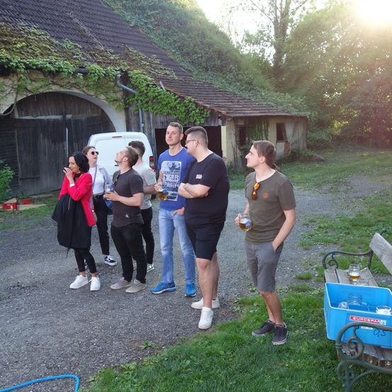 Eine Gruppe von Menschen steht draußen vor einem Gebäude mit einem von Moos bedeckten Dach. Sie unterhalten sich und trinken Bier.