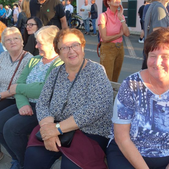 Eine Gruppe von Frauen sitzt auf einer Bank und lächelt, mit einer weiteren Frau im Hintergrund, die ein Telefon hält.