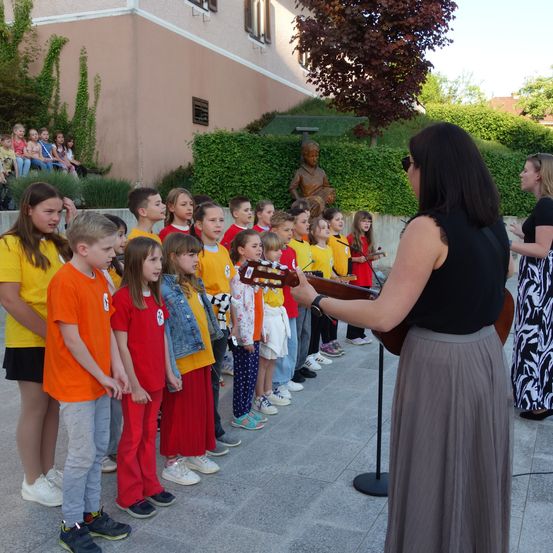 Eine Gruppe von Kindern in verschiedenen farbigen T-Shirts steht in einer Reihe, während eine Frau vor ihnen die Gitarre spielt. Dahinter steht eine Statue vor einem Gebäude mit einem Garten.
