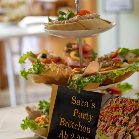 Ein dreistufiger Teller mit verschiedenen Sandwiches, die aufeinandergestapelt sind. Ein Schild für Saras Party Brotchen ist sichtbar.