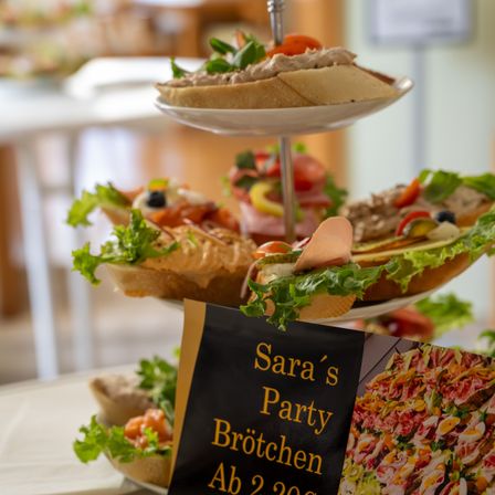 Ein dreistufiger Teller mit verschiedenen Sandwiches, die aufeinandergestapelt sind. Ein Schild für Saras Party Brotchen ist sichtbar.