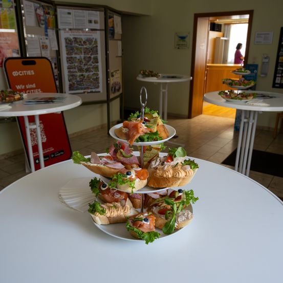 Ein weißer Tisch hat einen dreistufigen Stand mit belegtem Brot. Dahinter befinden sich weitere Tische mit Essen. Poster und ein roter Stand mit dem Wort 'Städte' sind auf der linken Seite. Eine Person ist in der Nähe der Tür.