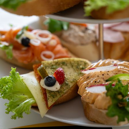 Ein dreistufiger Ständer hält verschiedene Sandwiches, darunter eines mit Räucherlachs, Käse und Salat, ein weiteres mit cremiger Aufstrich und Radieschen und ein drittes mit einem Käsestück, eingelegten Gurken und einer Erdbeere.