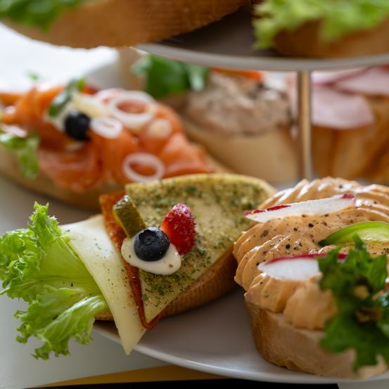 Ein dreistufiger Ständer hält verschiedene Sandwiches, darunter eines mit Räucherlachs, Käse und Salat, ein weiteres mit cremiger Aufstrich und Radieschen und ein drittes mit einem Käsestück, eingelegten Gurken und einer Erdbeere.