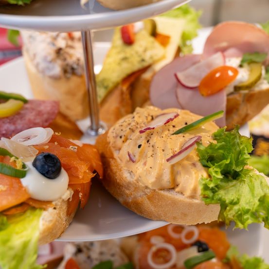 Ein dreischichtiges Gericht zeigt eine Vielzahl von Sandwiches, darunter Räucherlachs mit einer Blaubeere, Schinken mit Gurken und eine Auflauf mit grünem Gemüse.