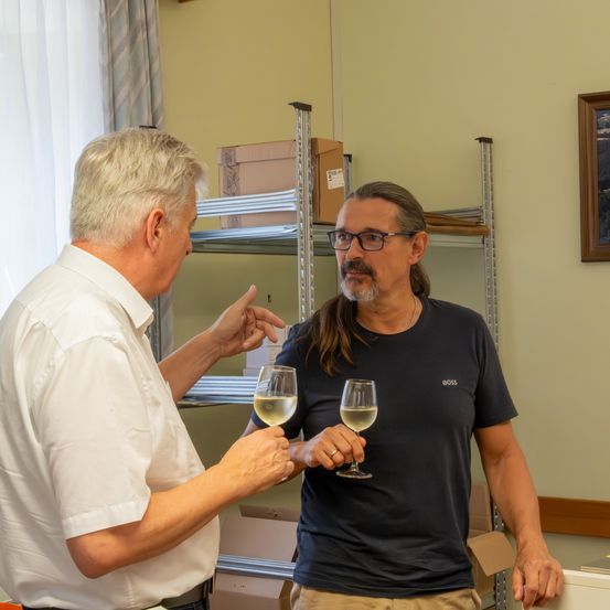 Zwei Männer in einem Raum mit Regalen im Hintergrund. Ein Mann hält ein Glas Weißwein, und der andere zeigt darauf.