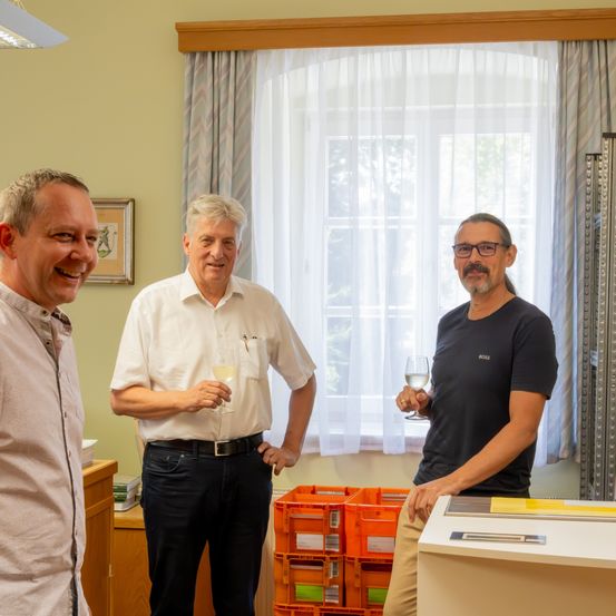 Drei Männer stehen in einem Raum, lächeln und halten Weingläser. Hinter ihnen ein Fenster mit weißen Vorhängen und einem gerahmten Bild an der Wand. In der Nähe orangefarbene Lagerkisten und eine weiße Maschine.