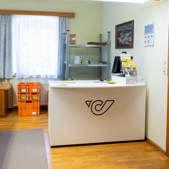 Ein Büroraum mit einem weißen Empfangstresen, einem Monitor, einem Kalender an der Wand und einem Fenster mit Vorhängen. Es gibt orangefarbene Lagerboxen und einen Heizkörper.