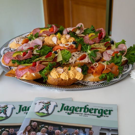 Ein Teller mit verschiedenen Sandwiches und Fleisch auf einem Tisch. Es gibt auch eine Zeitschrift mit dem Titel 'Jagerberger Gemeindenachrichten'.