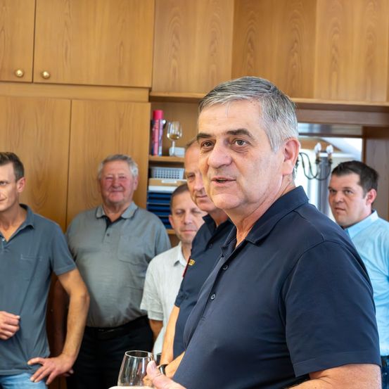 Eine Gruppe von Männern, darunter ein älterer Mann in einem dunkelblauen Poloshirt, steht in einem Raum mit Holzschränken und Regalen. Sie scheinen ein Gespräch zu führen.