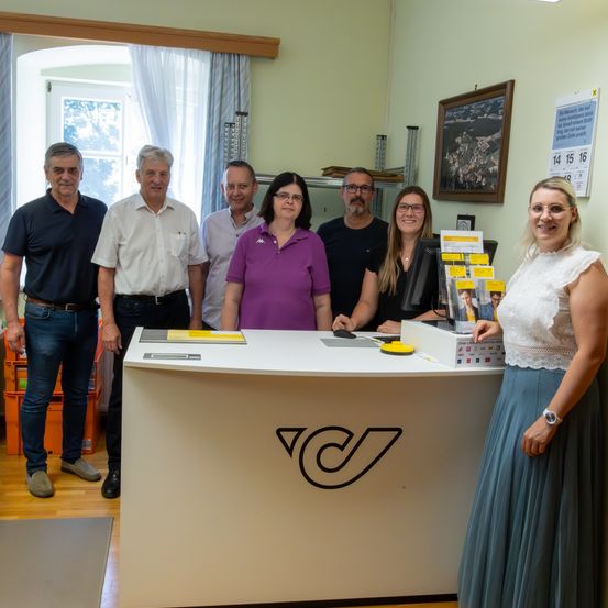 Eine Gruppe von Menschen steht in einem Büro mit einem Empfangstresen. Fünf Männer und eine Frau posieren für ein Foto. Ein Kalender ist an der Wand angeheftet.