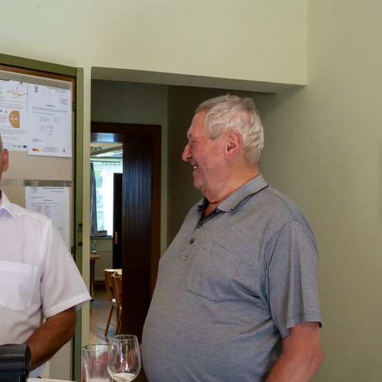 Ein älterer Mann in einem grauen Shirt steht lächelnd, möglicherweise im Gespräch mit einem Mann in einem weißen Shirt. Dahinter befindet sich eine Aushängetafel mit angehefteten Papieren. Im Hintergrund gibt es eine offene Tür, Stühle und einen Tisch.